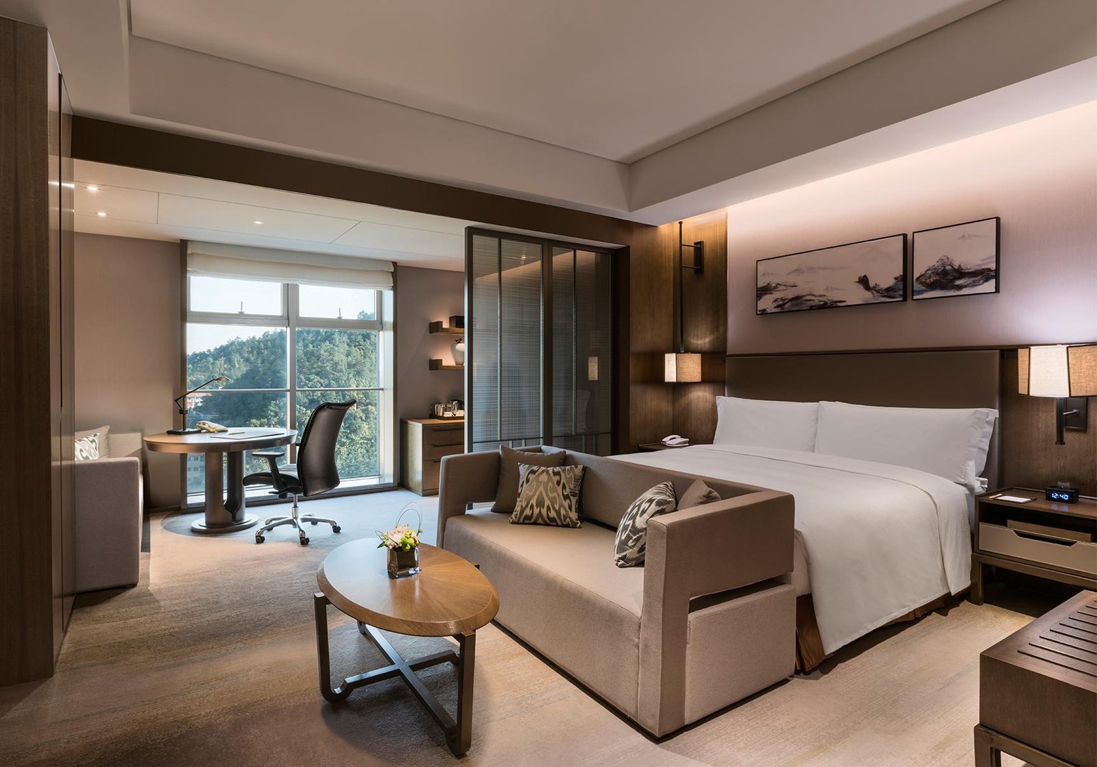 Hotel guest room at DoubleTree by Hilton Shiyan, Hubei, featuring a king-size bed, sofa, wooden coffee table, work desk, large windows overlooking greenery, and warm contemporary decor.