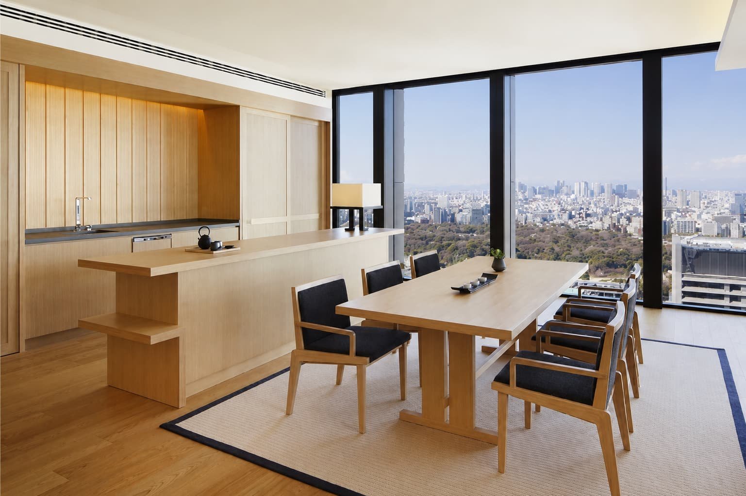 Hotel dining and kitchen area at Atami Hotel, Japan, featuring a light wood kitchen, dining table with upholstered chairs, and floor-to-ceiling windows overlooking the city and surrounding landscape.