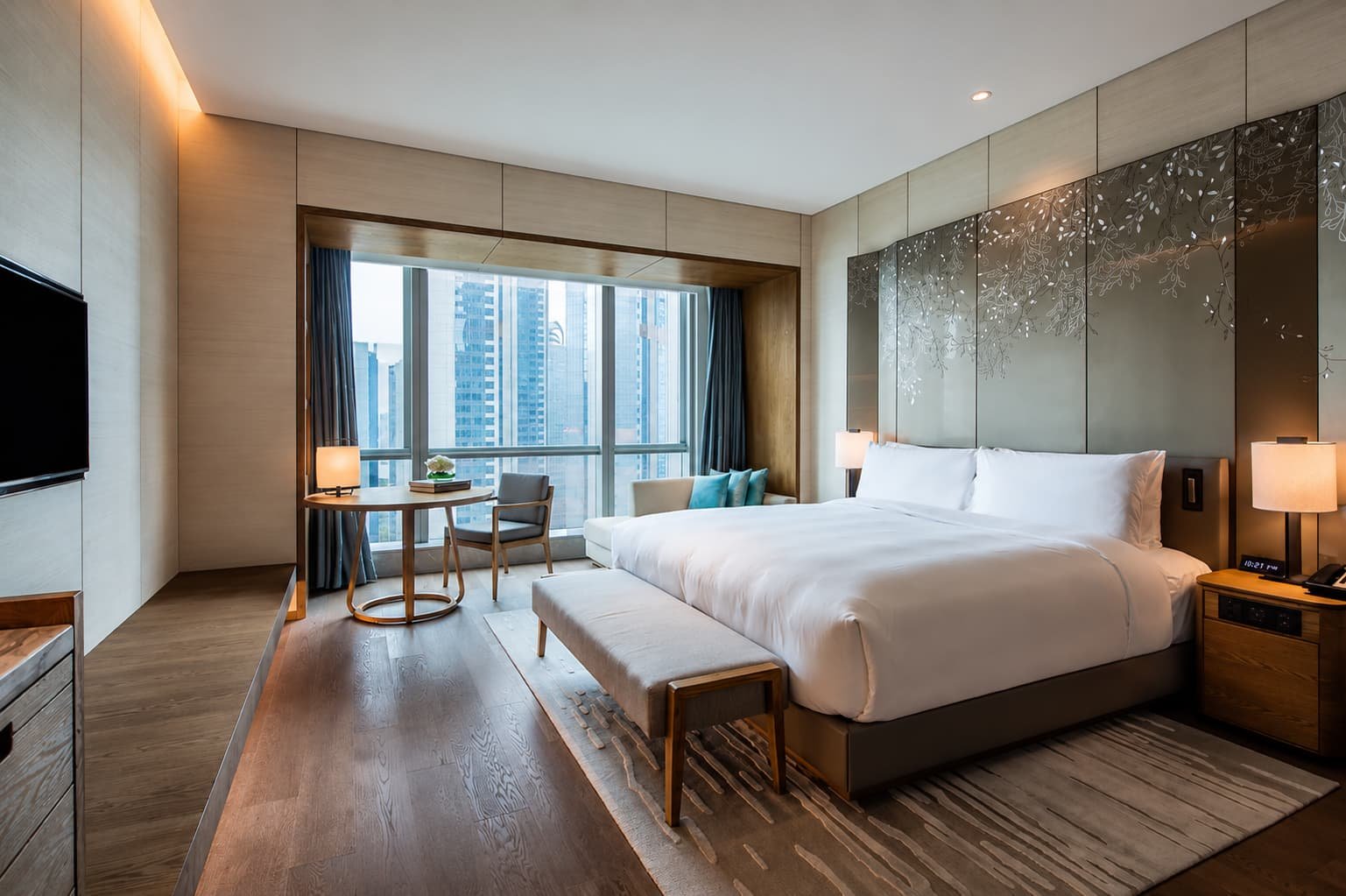 Modern guest room at Conrad Guangzhou Hotel, featuring a king-size bed with white linens, a patterned headboard, upholstered bench, wooden furnishings, and floor-to-ceiling windows overlooking the city skyline.