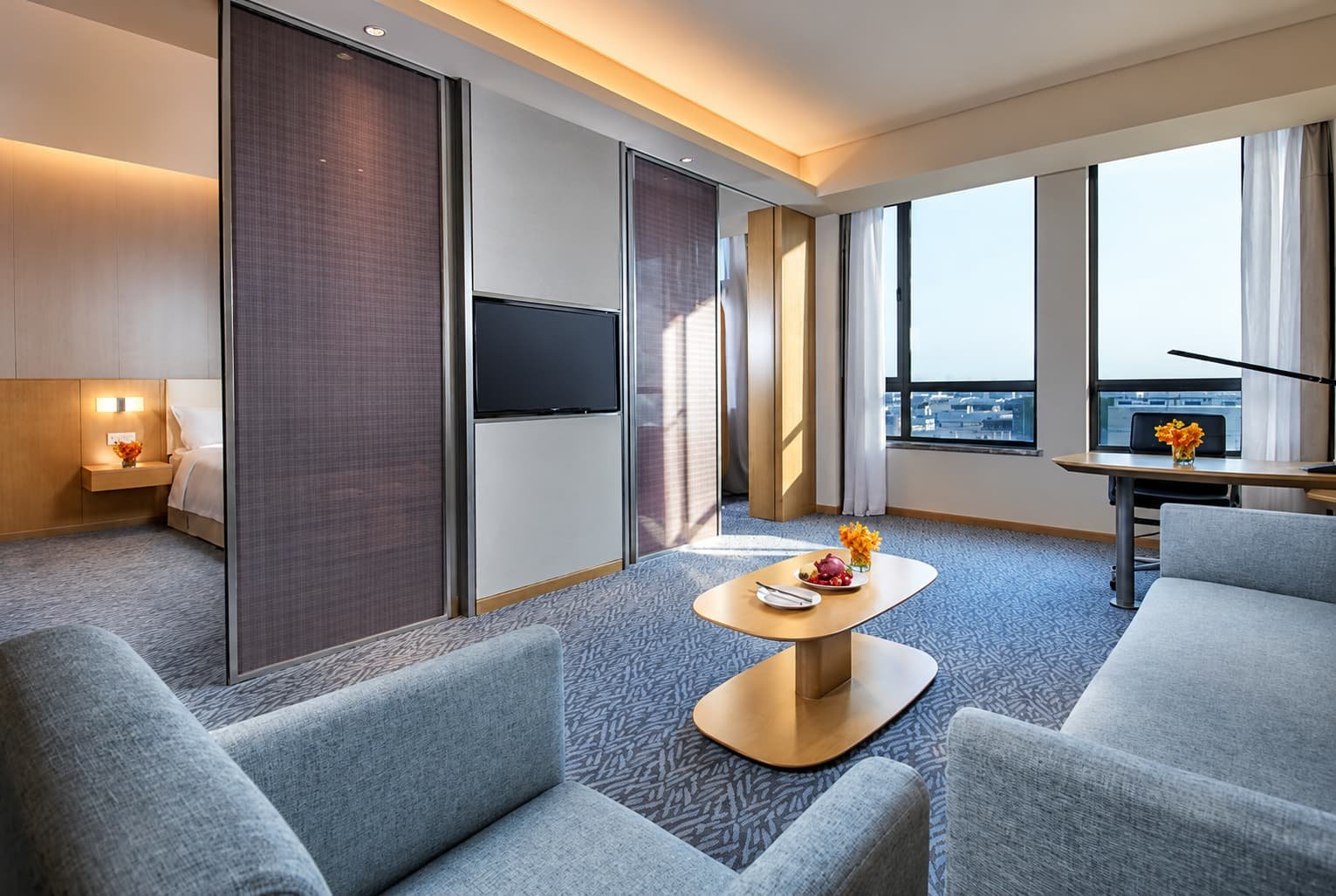 Modern hotel suite at Holiday Inn Express Nantong, featuring a bedroom, spacious sitting area with upholstered sofas, a wooden coffee table, wall-mounted TV, work desk, and large windows overlooking the city.
