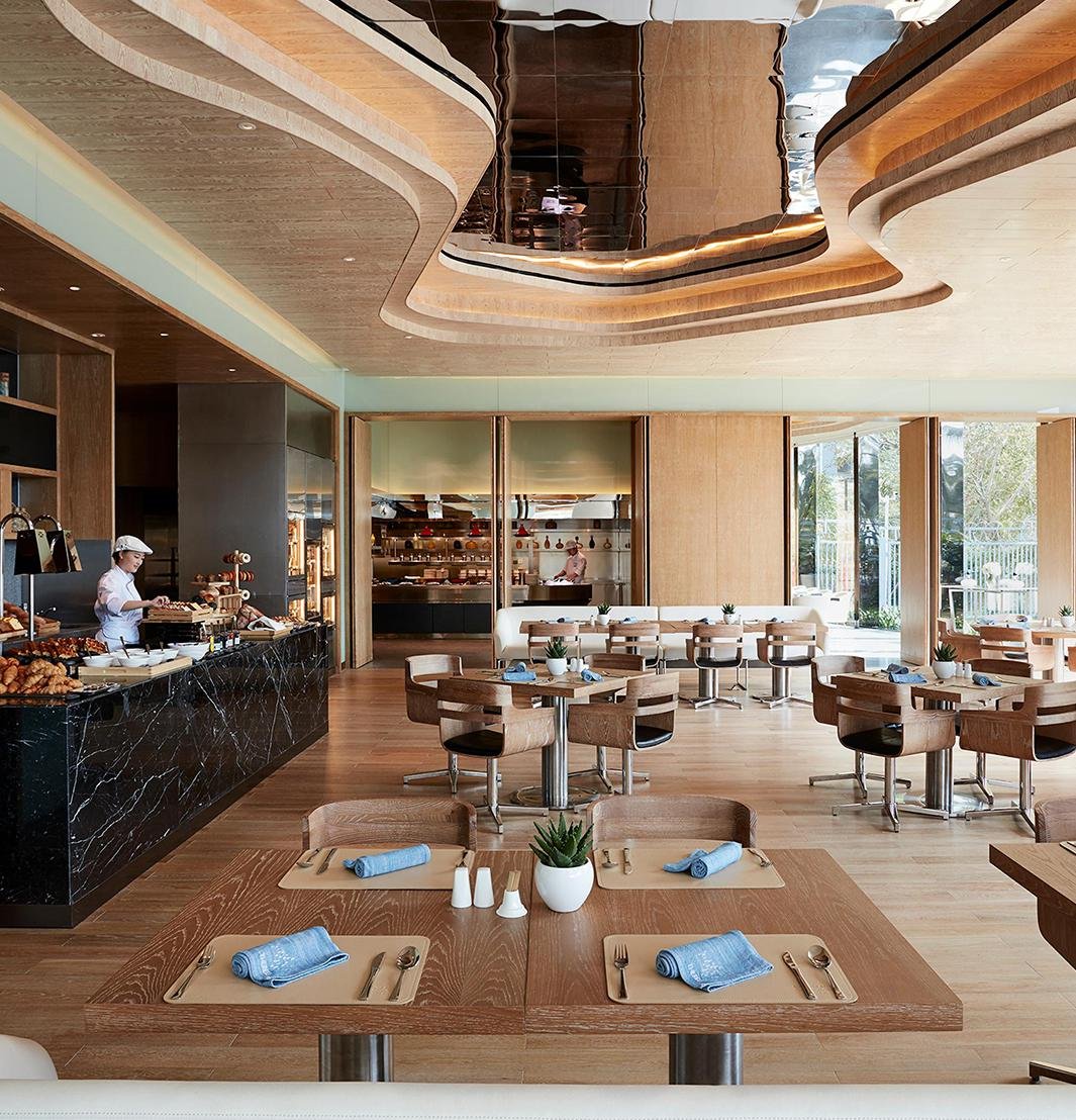 estaurant dining area at Shenzhen Dapeng Marriott Hotel, featuring wooden tables and chairs, neatly set place settings with blue napkins, a marble service counter, and a modern curved wood-and-metal ceiling.